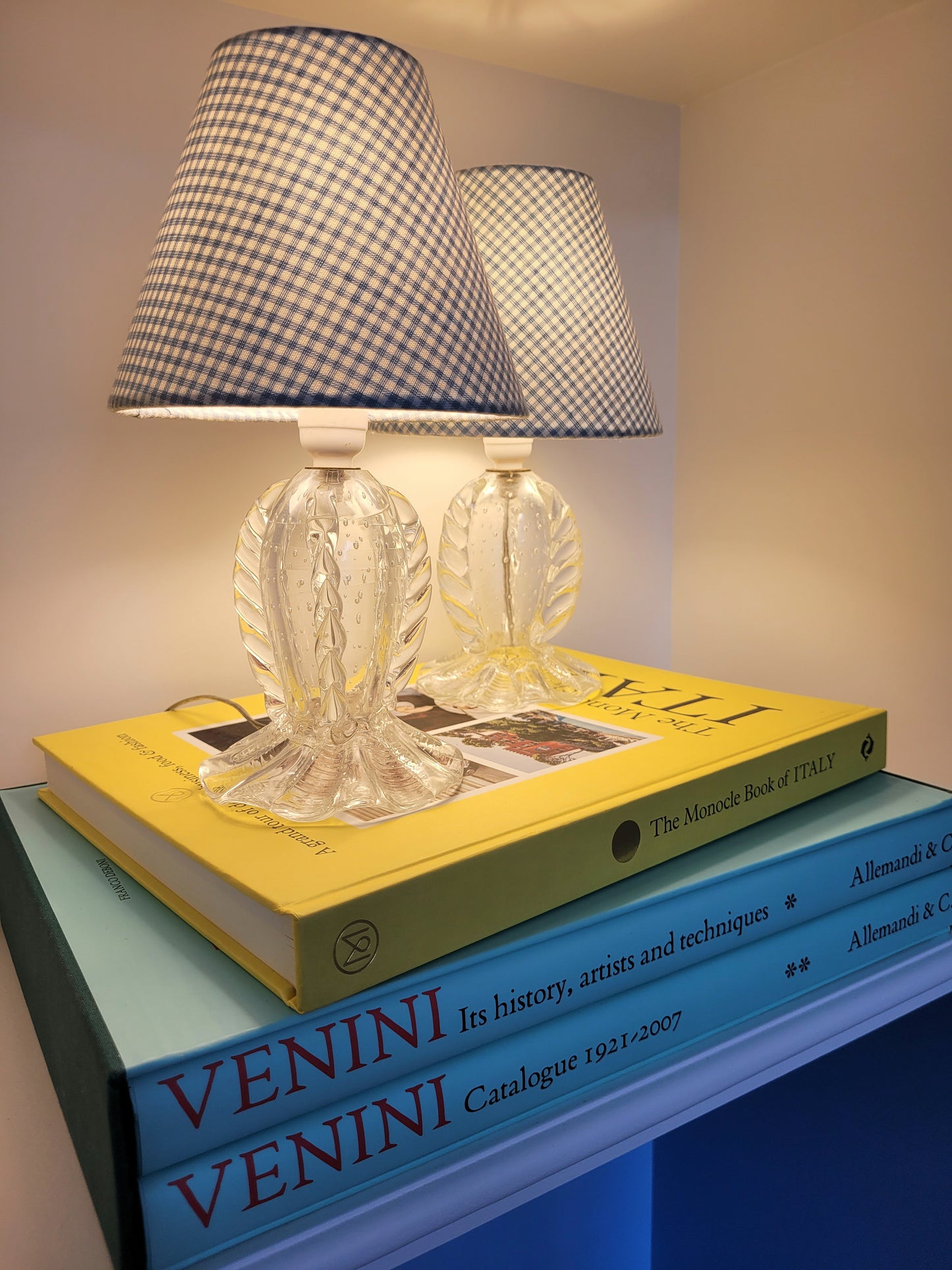 side view of a rare pair of vintage clear Murano table lamps with shades stacked on books illuminated