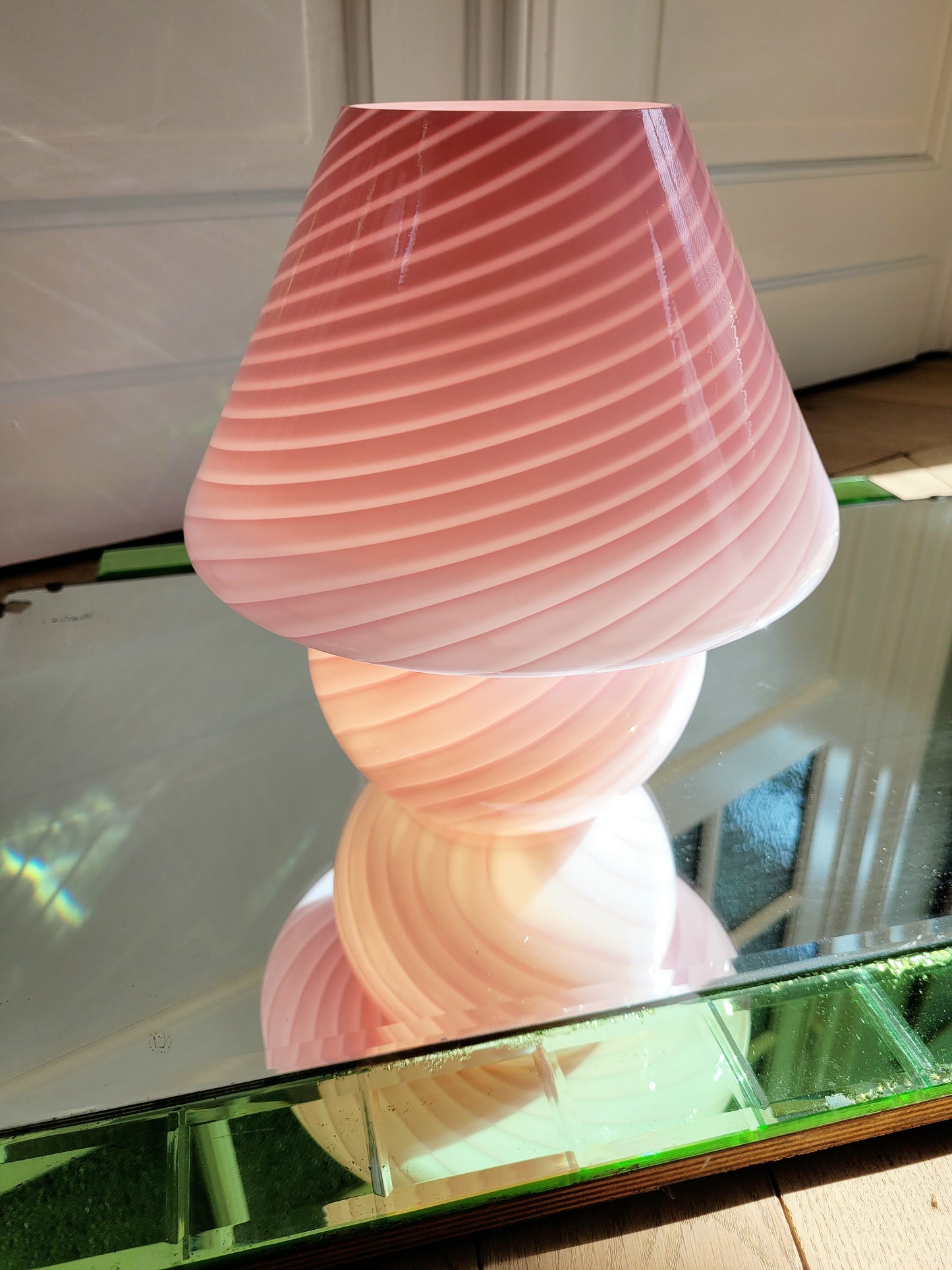 front view of a pink and white swirl Murano mushroom table lamp on a mirror with green edging. The lamp is illuminated.