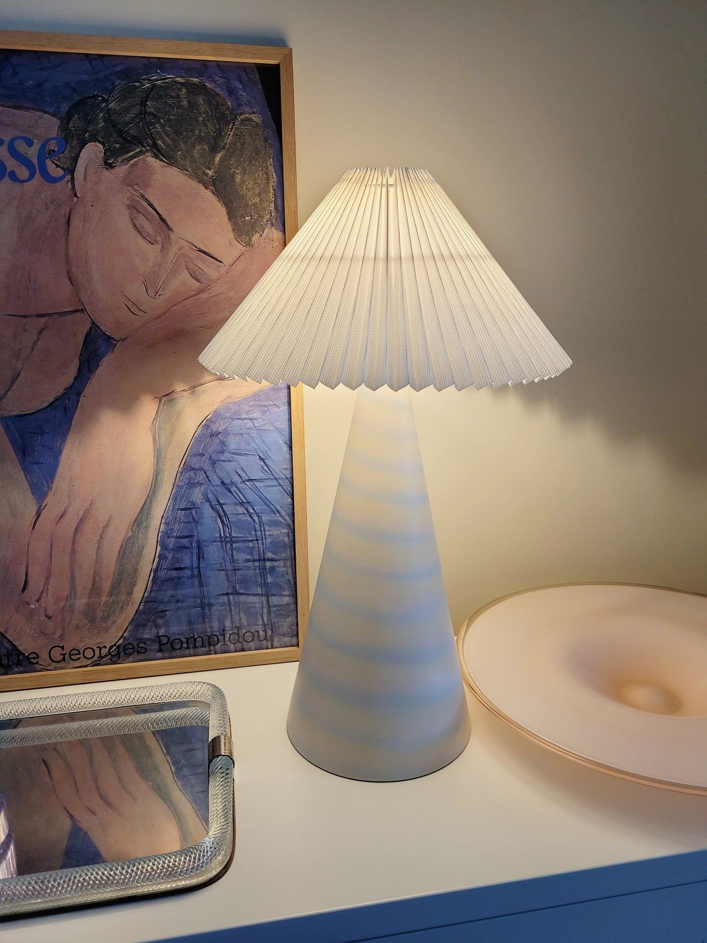 detail of an illuminated pastel ceramic table lamp with white pleated lampshade on a cabinet with Italian vintage glass pieces and a vintage Matisse exhibition poster