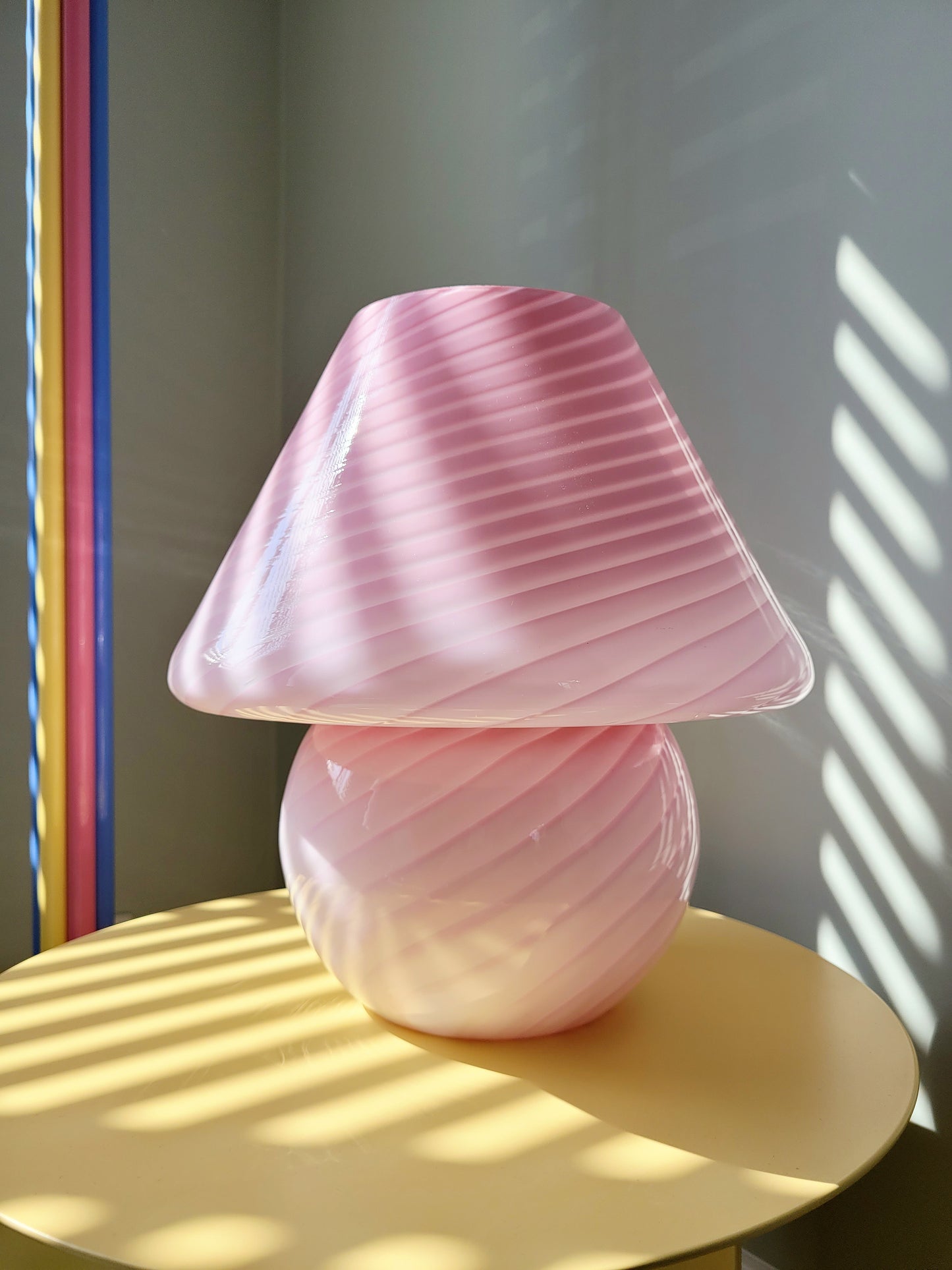 front view of a pink and white swirl Murano mushroom table lamp on a yellow side table. Rays of sunshine flooding in.