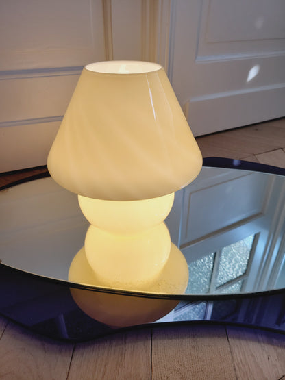front view of an ochre yellow swirl Murano mushroom table lamp on a mirror with cobalt blue edging. The lamp is illuminated.