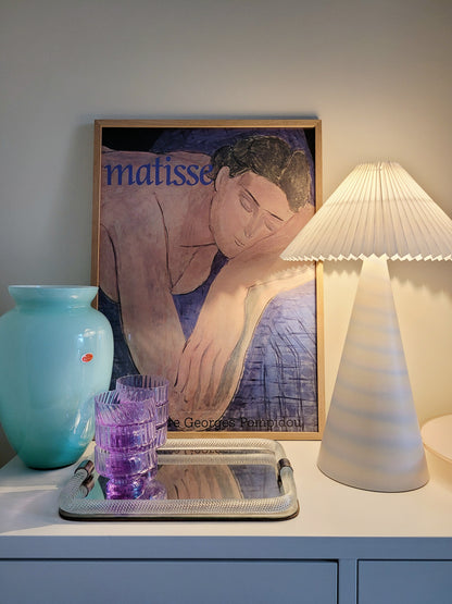 front view of an illuminated pastel ceramic table lamp with white pleated lampshade on a cabinet with Italian vintage glass pieces and a vintage Matisse exhibition poster