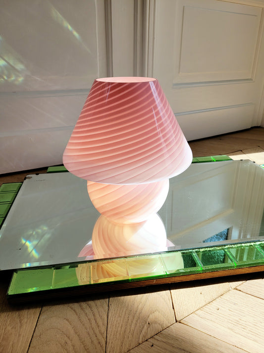 front view of a pink and white swirl Murano mushroom table lamp on a mirror with green edging. The lamp is illuminated.