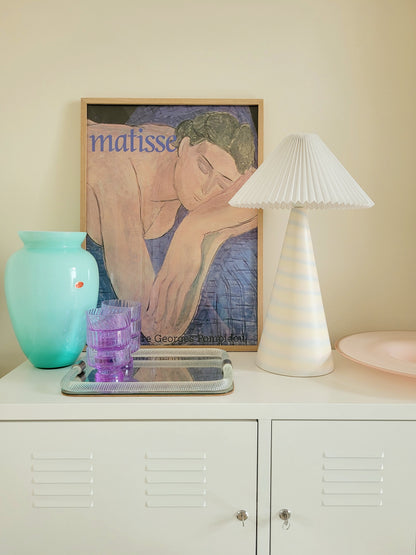 front view of a pastel ceramic table lamp with white pleated lampshade on a cabinet with Italian vintage glass pieces and a vintage Matisse exhibition poster