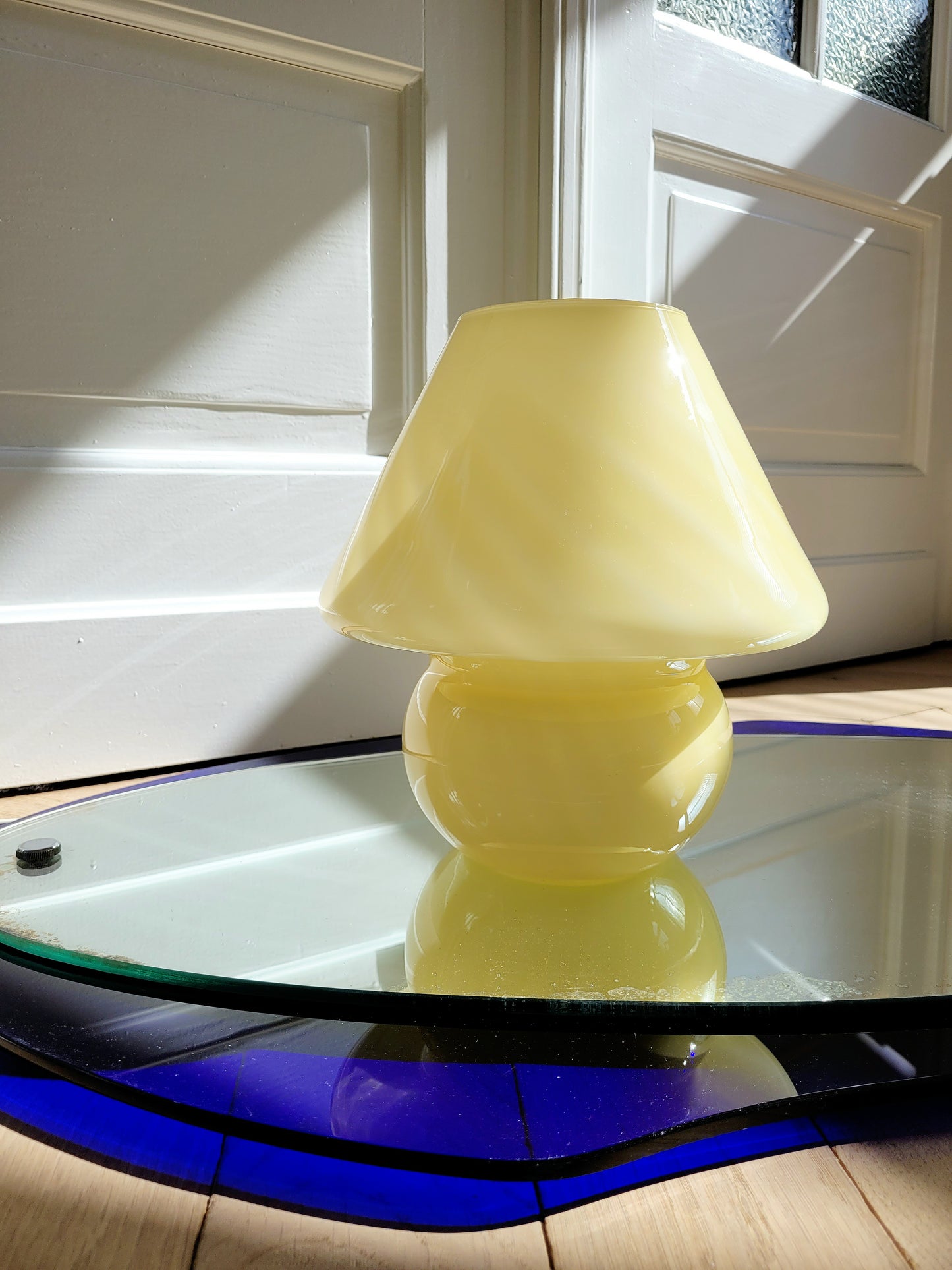 front view of an ochre yellow swirl Murano mushroom table lamp on a mirror with cobalt blue edging. 