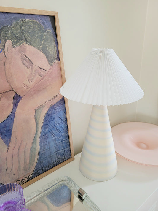 detail of a pastel ceramic table lamp with white pleated lampshade on a cabinet with Italian vintage glass pieces and a vintage Matisse exhibition poster