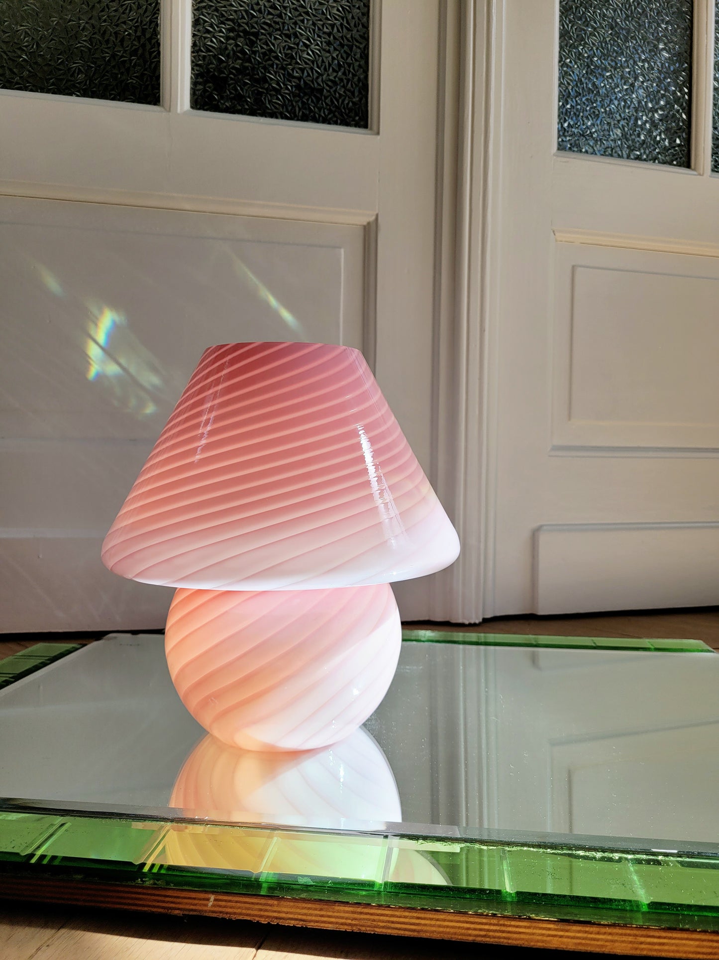 front view of a pink and white swirl Murano mushroom table lamp on a mirror with green edging. The lamp is illuminated.