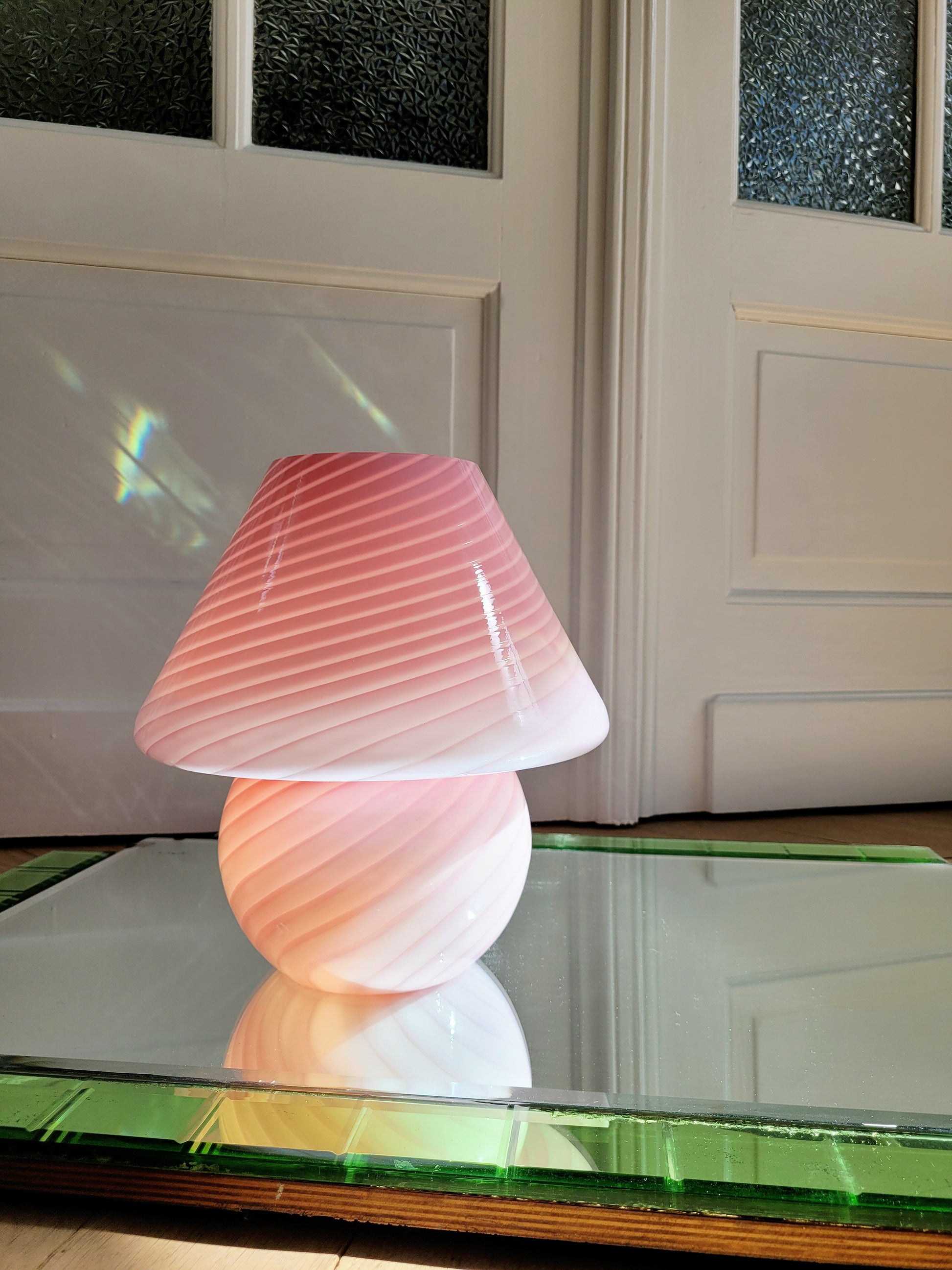 front view of a pink and white swirl Murano mushroom table lamp on a mirror with green edging. The lamp is illuminated.