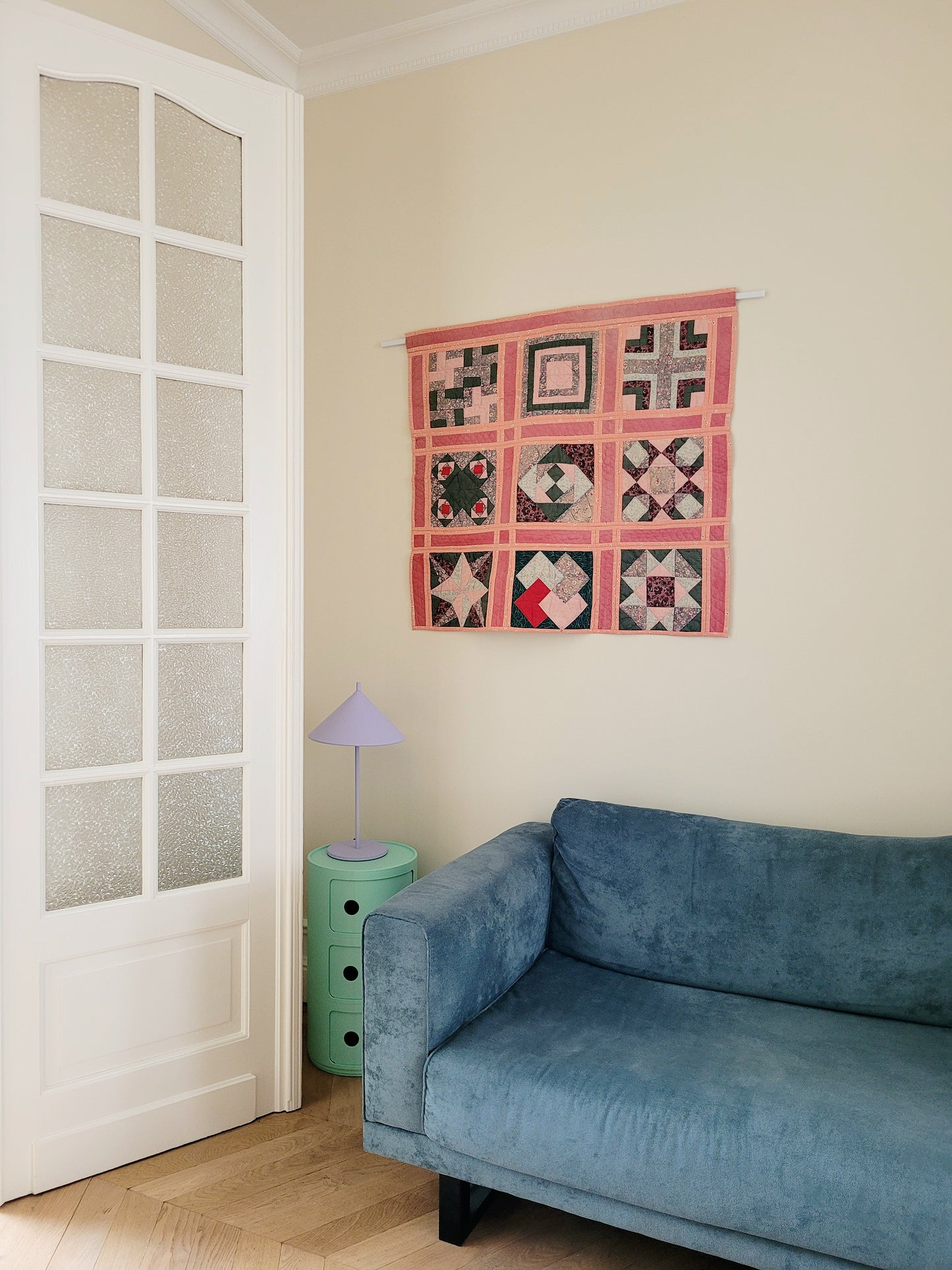 side view of a pink and green vintage handmade patchwork quilt with unique geometric pattern against a yellow wall and above a green side table with purple table lamp and teal sofa