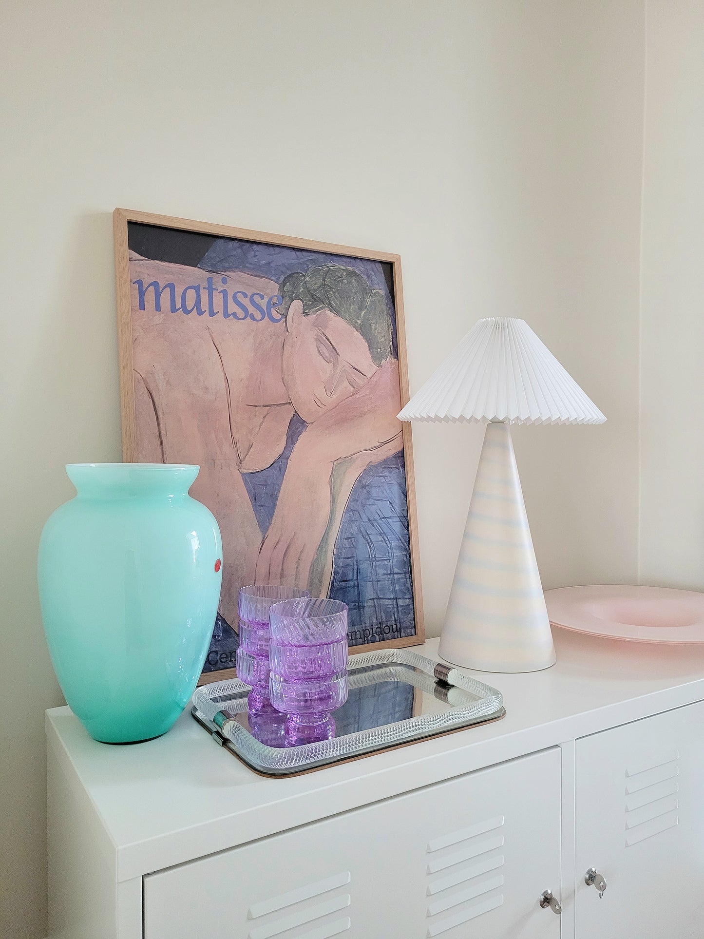 side view of a pastel ceramic table lamp with white pleated lampshade on a cabinet with Italian vintage glass pieces and a vintage Matisse exhibition poster