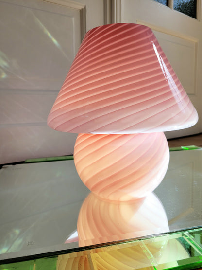 detail of a pink and white swirl Murano mushroom table lamp on a mirror with green edging. The lamp is illuminated.
