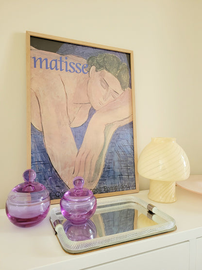 front view of two vintage clear purple Murano bonbonnieres on a Murano mirror tray with a Matisse exhibition poster end yellow Murano table lamp in the background