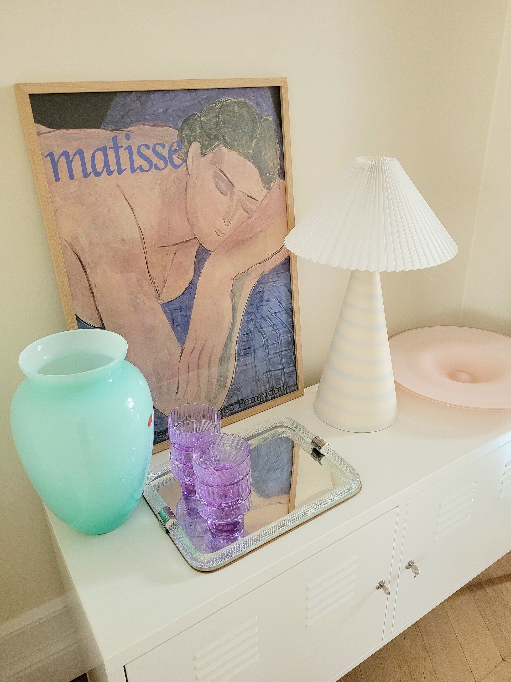side view of a pastel ceramic table lamp with white pleated lampshade on a cabinet with Italian vintage glass pieces and a vintage Matisse exhibition poster