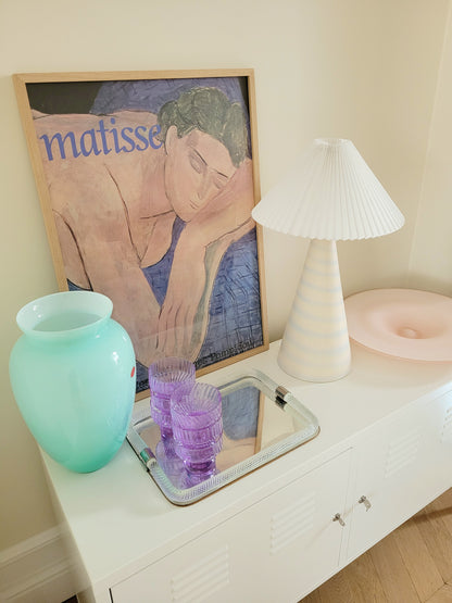 side view of a pastel ceramic table lamp with white pleated lampshade on a cabinet with Italian vintage glass pieces and a vintage Matisse exhibition poster