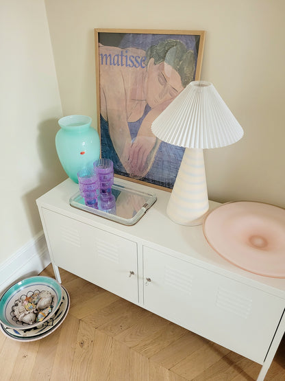side view of a pastel ceramic table lamp with white pleated lampshade on a cabinet with Italian vintage glass pieces and a vintage Matisse exhibition poster