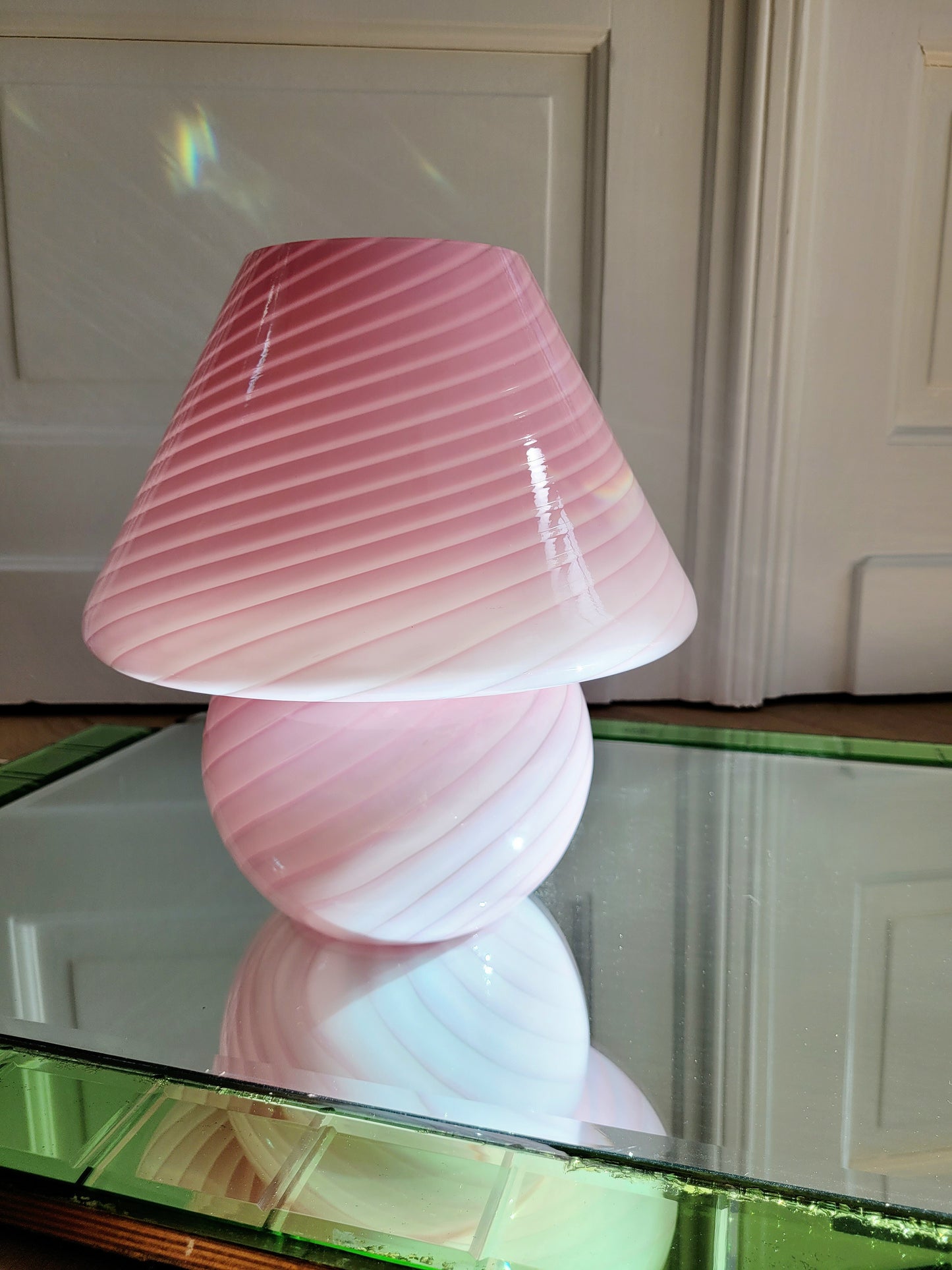 front view of a pink and white swirl Murano mushroom table lamp on a mirror with green edging. 