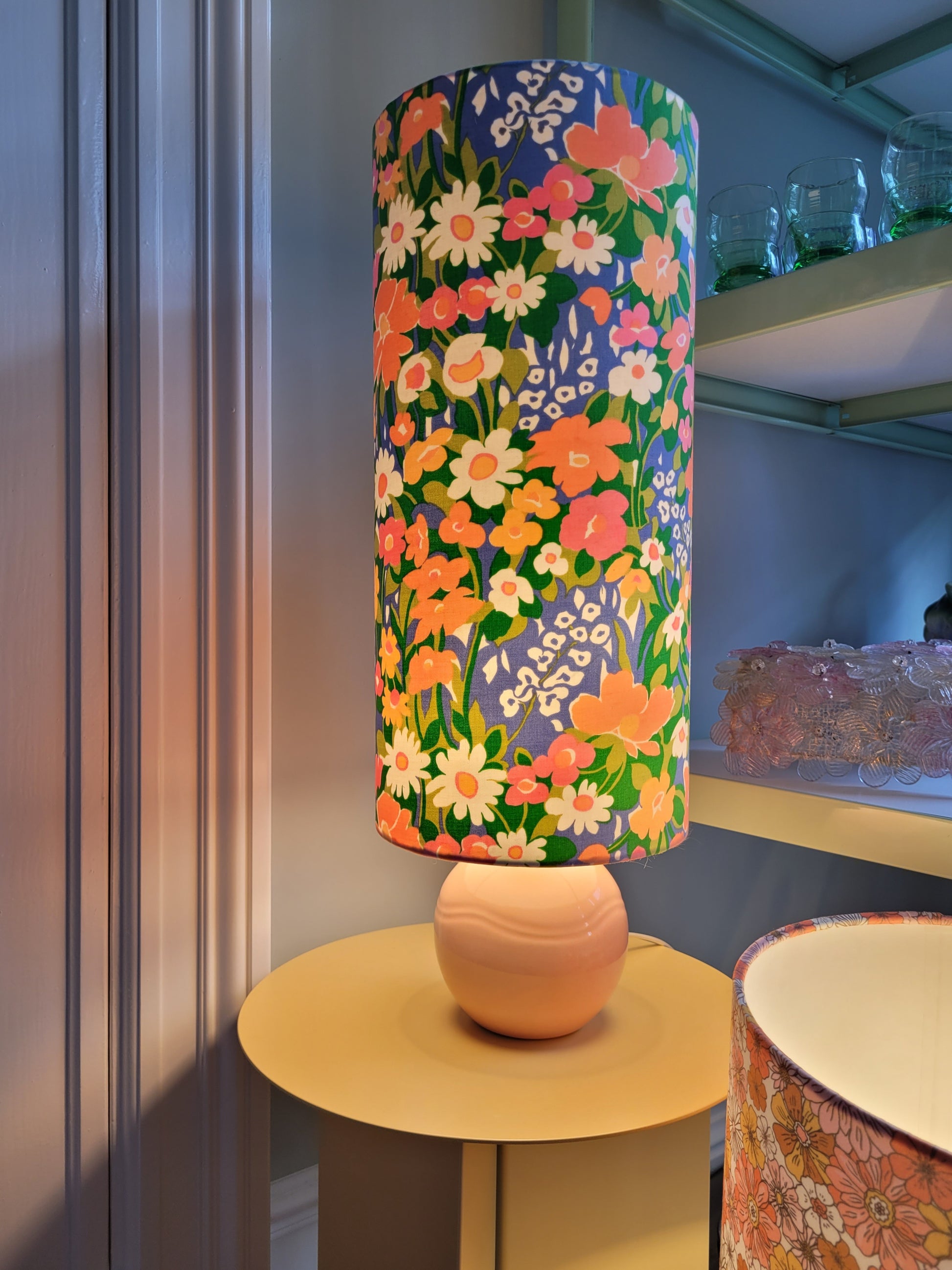 A cheerful table lamp with a pink/coral ceramic base paired with an extra-tall, cylindrical fabric lampshade in a lively retro floral print from the 80s.