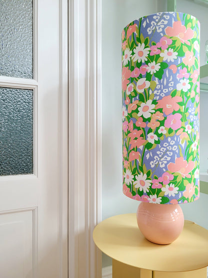 front view of a cheerful table lamp with a pink/coral ceramic base paired with an extra-tall, cylindrical fabric lampshade in a lively retro floral print from the 80s.