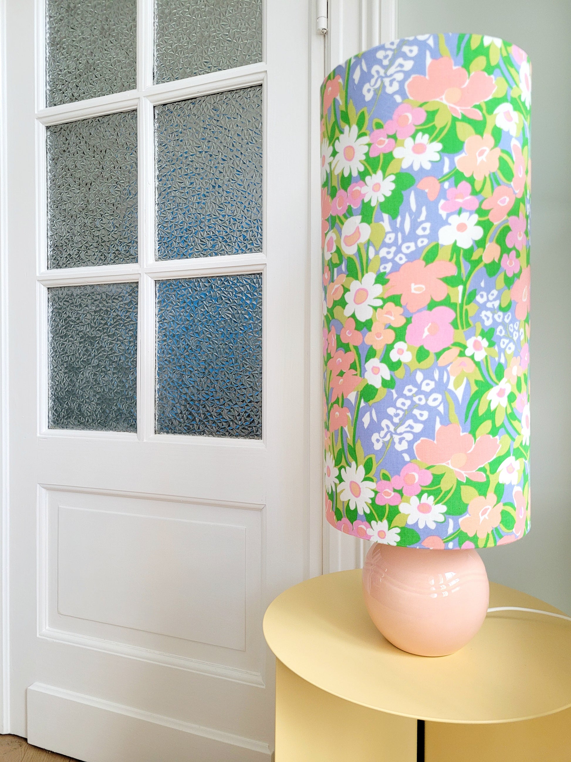 A cheerful table lamp with a pink/coral ceramic base paired with an extra-tall, cylindrical fabric lampshade in a lively retro floral print from the 80s.